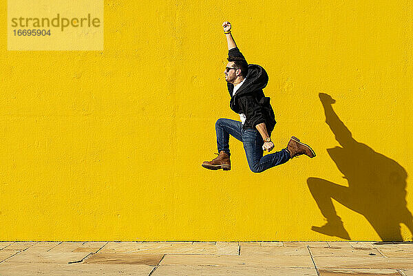 Junger Mann mit Sonnenbrille  der vor eine gelbe Wand springt.