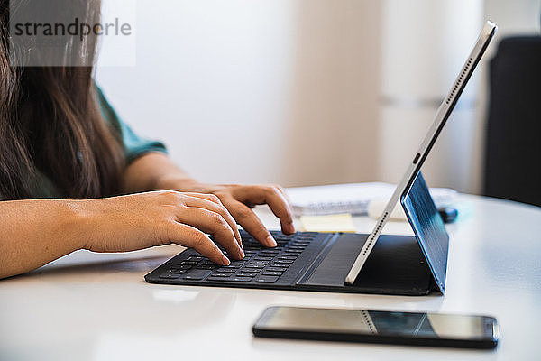 Nahaufnahme der Hände einer Frau  die auf der Tastatur ihres Hybridgeräts aus Tablet und Laptop tippt  neben einem Telefon auf einem Tisch  während sie telearbeitet und sich an die neue Normalität nach der Coronavirus-Krise anpasst
