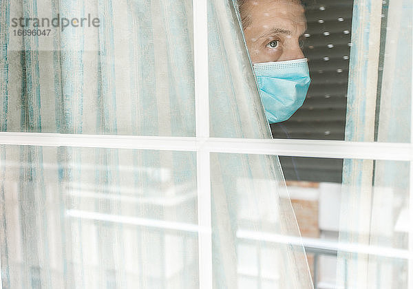 Foto eines weißen Mannes mittleren Alters mit medizinischer Gesichtsmaske  der während der Coronavirus-Quarantäne aus dem Fenster schaut