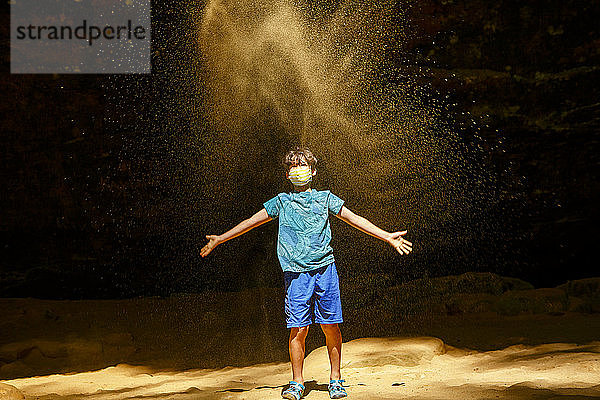 Ein Junge mit Gesichtsmaske steht in einer sonnenbeschienenen Schlucht und wirft goldenen Sand auf