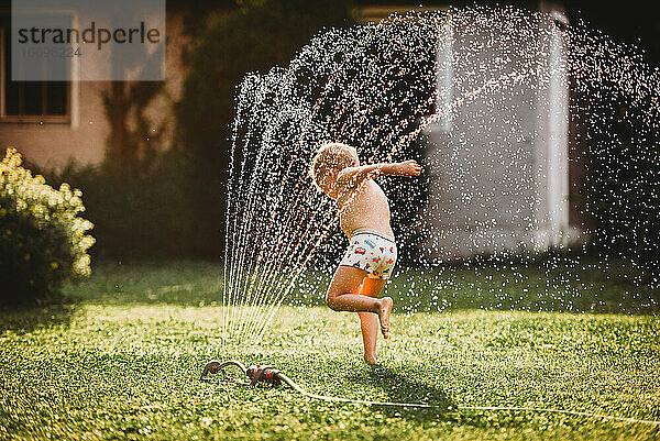 Junge weiße Junge läuft unter dem Wasser aus dem Sprinkler im Garten