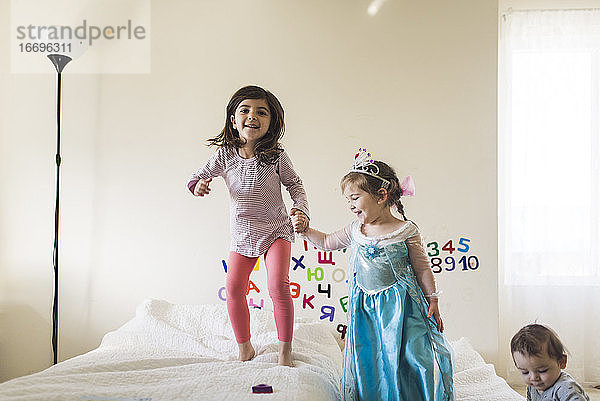 Lachendes 6-jähriges Mädchen und 4-jähriges Mädchen im Prinzessinnenkleid springen auf dem Bett