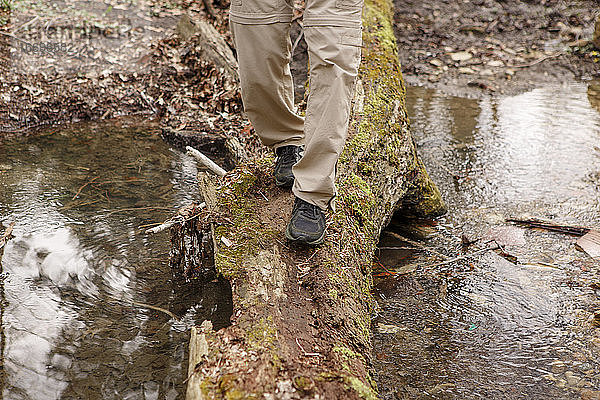 Untere Hälfte eines Mannes beim Überqueren eines Baches auf einem moosbewachsenen Baumstamm in der Natur