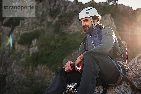 Konzept: Abenteuer. Mann mit Helm  Klettergurt und Rucksack. Nachdenklich auf einem Felsen sitzend. Solare Leuchterscheinung.