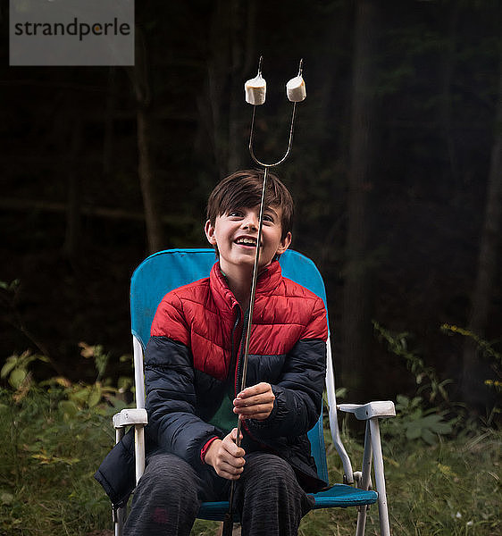 Fröhlicher Junge  der geröstete Marshmallows an einem Metallstab betrachtet.