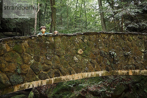Zwei kleine Kinder spähen mit ihren Köpfen über eine Steinbrücke im Wald