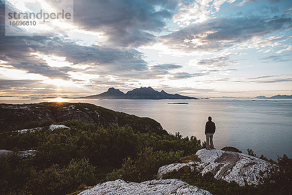 Mann steht auf einem Felsen bei Sonnenuntergang am Meer und schaut auf eine Insel
