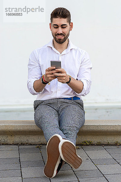 Junger bärtiger Mann sitzt an einer weißen Wand und benutzt ein Mobiltelefon