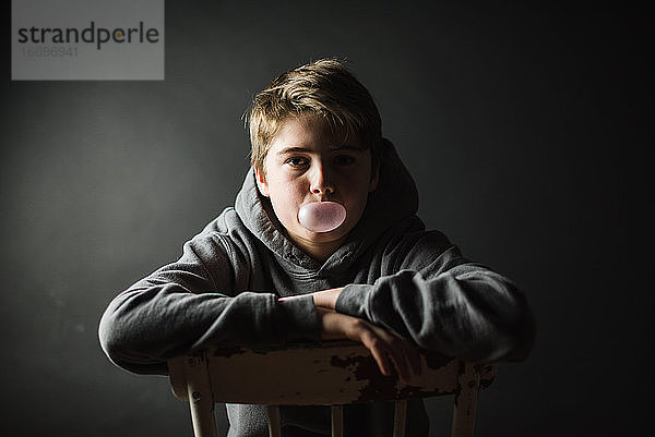 Teenager im Kapuzenpulli sitzt auf einem Stuhl in einem dunklen Raum und bläst eine Seifenblase.