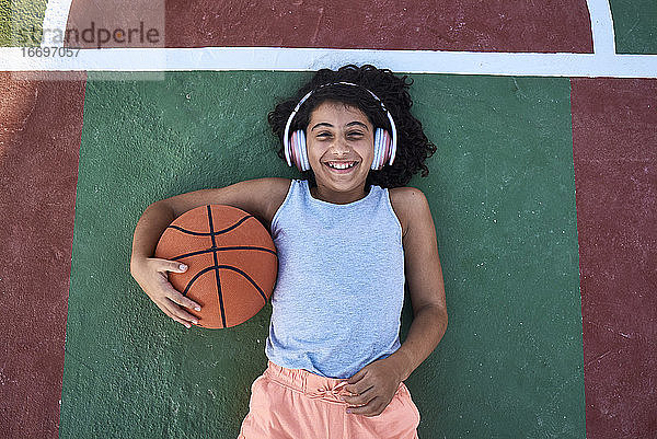Ein kleines Mädchen mit lockigem Haar liegt lachend auf einem Basketballplatz