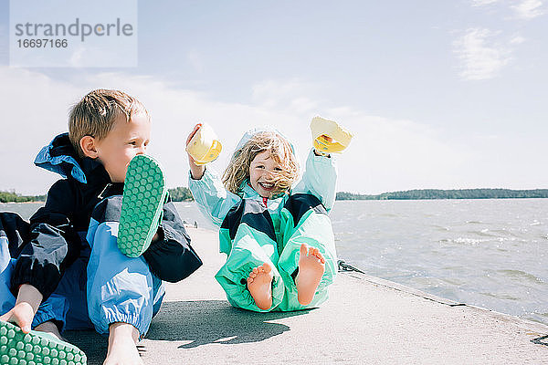 Geschwister  die an einem windigen Tag am Strand lachend ihre Schuhe festhalten