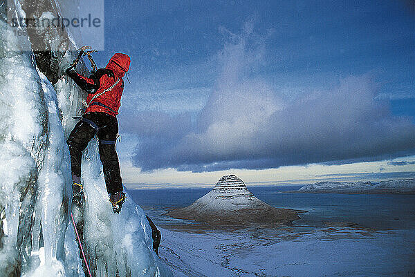 Mann erklimmt gefrorenen Wasserfall über dem Grundafjordur-Fjord in Island