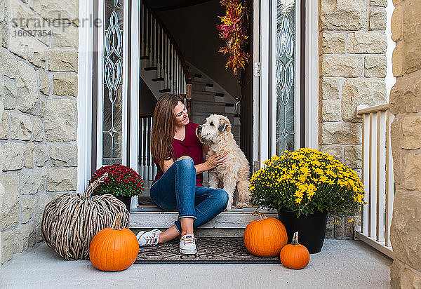 Frau und ihr Hund sitzen in der Tür eines herbstlich dekorierten Hauses.