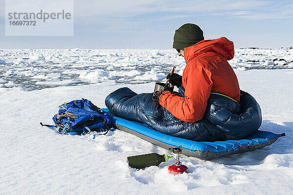 Männlicher Wanderer  der im Schlafsack auf einem schneebedeckten Berg sitzt und isst