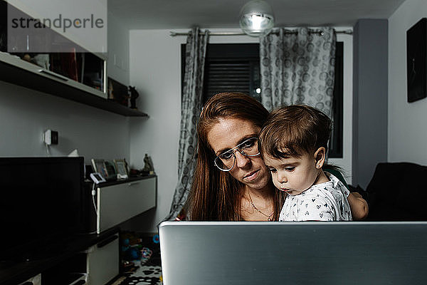 Frau mit kleinem Kind auf dem Arm benutzt Laptop zu Hause