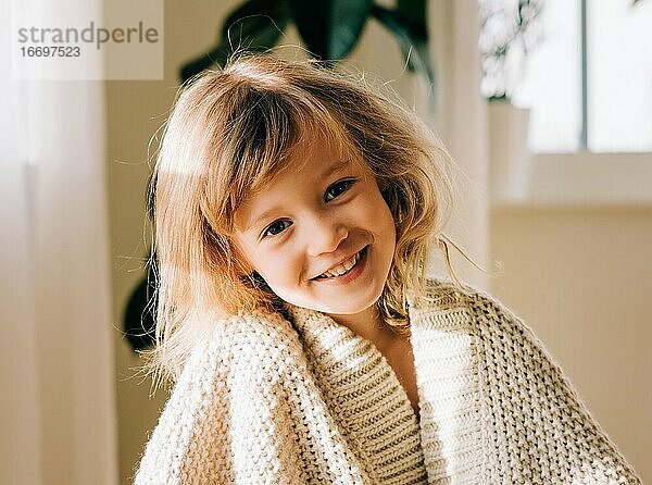 Porträt eines lächelnden jungen Mädchens  das zu Hause in eine Decke eingewickelt sitzt