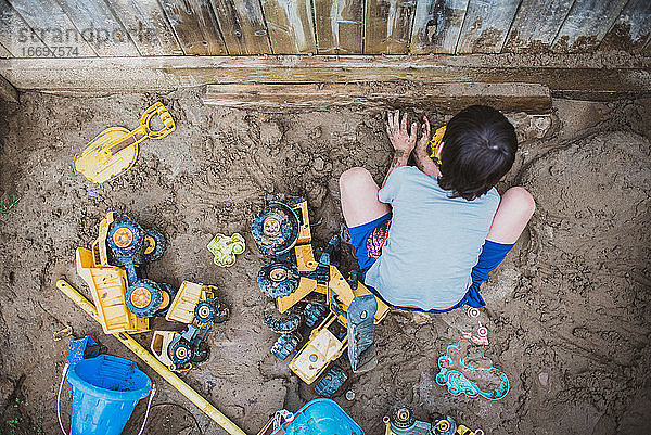 Draufsicht auf einen kleinen Jungen  der in einem schlammigen Sandkasten voller Spielzeug spielt.
