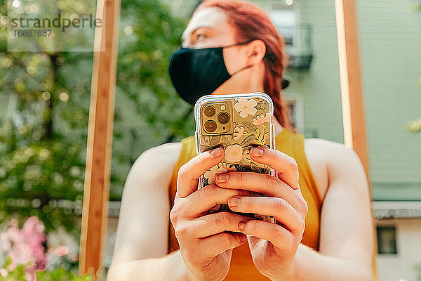 Junge Frau sitzt im Freien und schreibt eine SMS  sie trägt eine Gesichtsmaske.