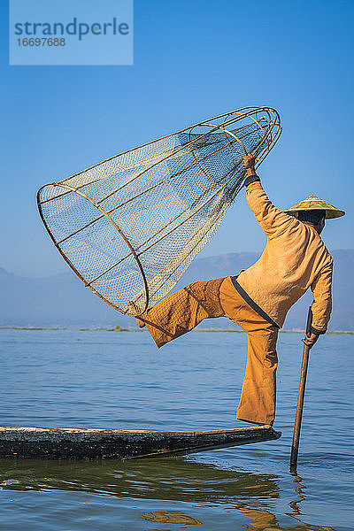 Rückansicht eines Intha-Fischers  der mit dem typischen konischen Fischernetz auf einem Boot vor dem klaren blauen Himmel posiert  Inle-See  Nyaungshwe  Myanmar