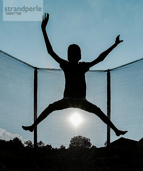 Vertikales Gegenlichtfoto einer Silhouette eines barfuß springenden oder fliegenden Jungen auf einem Trampolin mit Netz und der Sonne im Hintergrund  die Sonnenstrahlen auf seinem Schatten reflektiert