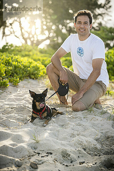 Junger fitter Mann hockt am Strand und kleiner schwarzer Hund leckt Nase