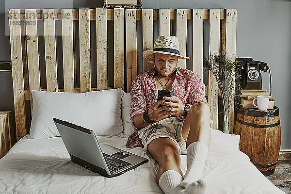 ein Mann  der mit seinem Handy und Laptop in einem Hotelbett arbeitet.