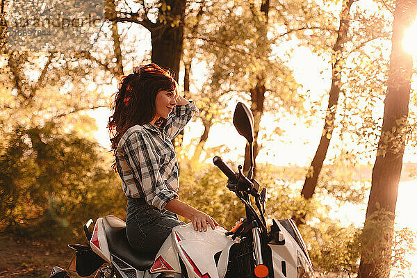 Glückliche junge Frau entspannt auf einem Motorrad sitzend inmitten von Bäumen