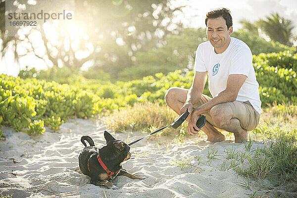 Junger fitter Mann hockt am Strand mit seinem kleinen schwarzen Hund in Hawaii