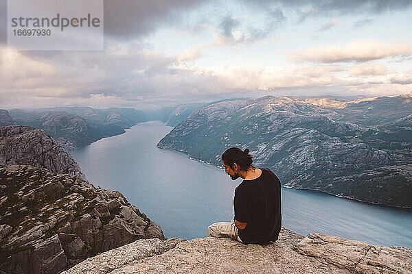 Mann schaut neugierig nach unten  sitzt in einem Felsen am Rande einer Klippe am Preikestolen  Norwegen