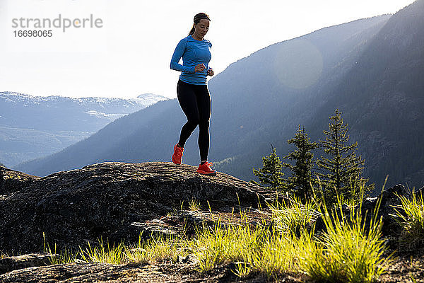 Vorderansicht einer Sportlerin in Sportkleidung  die an einem sonnigen Abend auf dem Lande auf Felsen in der Nähe von Bergen läuft