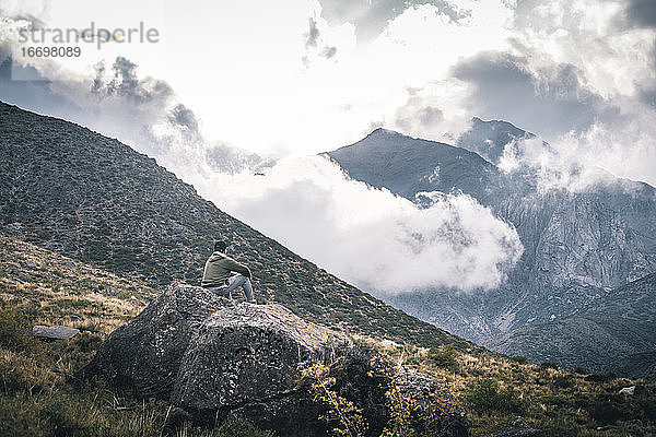junger Mann sitzt auf einem Felsen und bewundert Berge mit tief hängenden Wolken