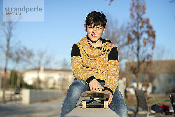 Junger Teenager sitzt auf seinem Skateboard auf einem Zaun und schaut in die Kamera