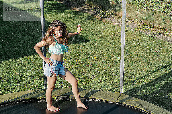 Horizontales Foto eines jungen brünetten Teenagers auf einem Trampolin  der sich mit der Hand auf das Netz in einem Park stützt. Das Mädchen trägt kurze Jeans und ein Top und sieht müde  passiv und ohne Energie aus.