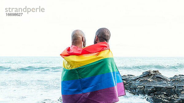 Lgbt Stolz Konzept. Homosexuelles Paar  das sich liebevoll umarmt  eingewickelt in die Regenbogenflagge mit Blick auf die Meereslandschaft.