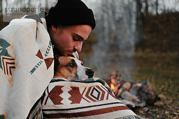 Mann in Ponchodecke am Lagerfeuer umarmt seinen Hund  kühler Herbst