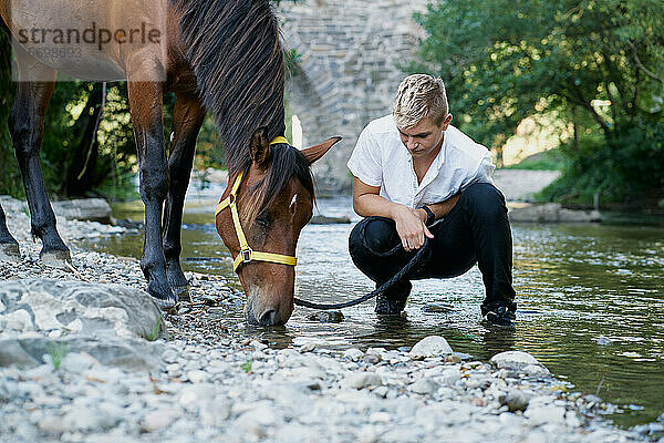 Porträt eines blonden jungen Mannes mit einem Pferd an einem Fluss