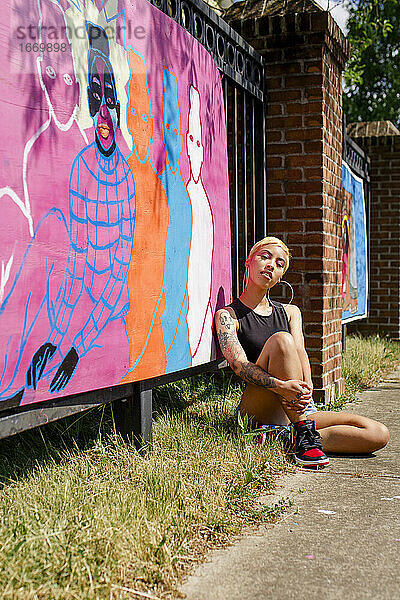 Eine schöne junge Künstlerin sitzt auf dem Boden und zeigt stolz ihr Wandgemälde