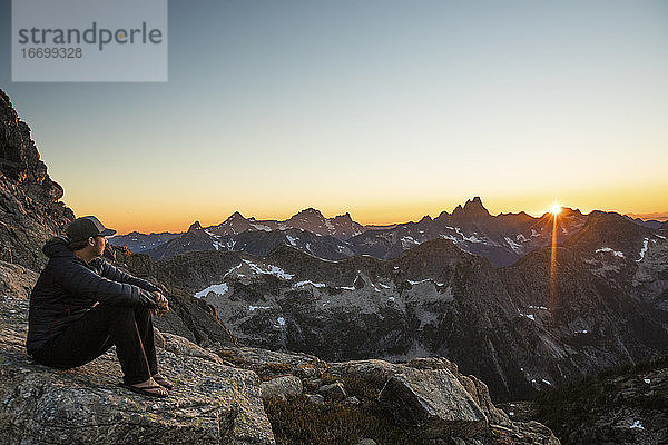 Aktiver  fitter Mann ruht sich auf einem Felsen in den Bergen aus und beobachtet den Sonnenuntergang.