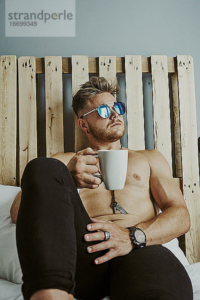 Ein blonder Mann mit blauer Sonnenbrille trinkt Kaffee im Bett