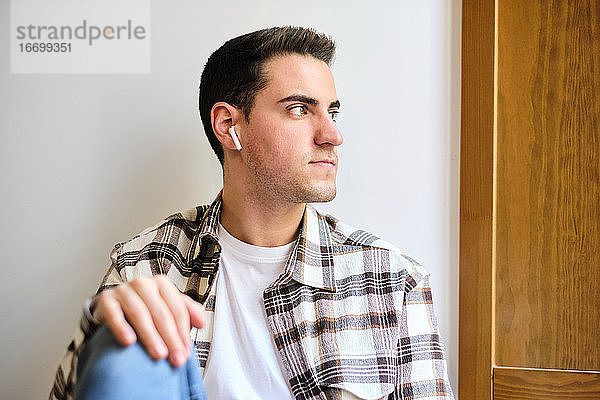 Porträt eines jungen Mannes  der zu Hause chillt und wegschaut  während er mit Kopfhörern Musik hört.