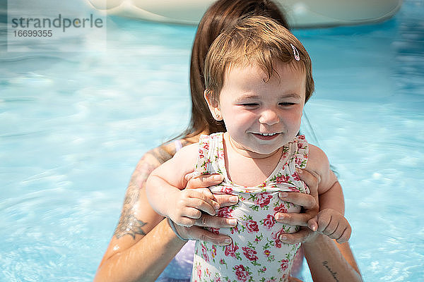 Kleines Mädchen lächelnd mit den Händen einer Frau in der Mitte eines Pools