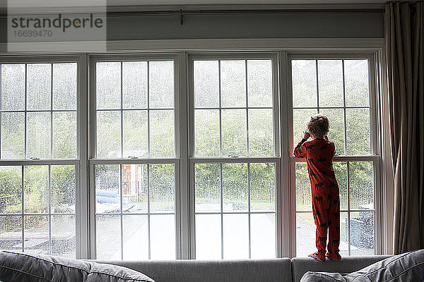 Wide Rear View of Boy in Red Pajamas Blick aus dem Fenster bei Regen