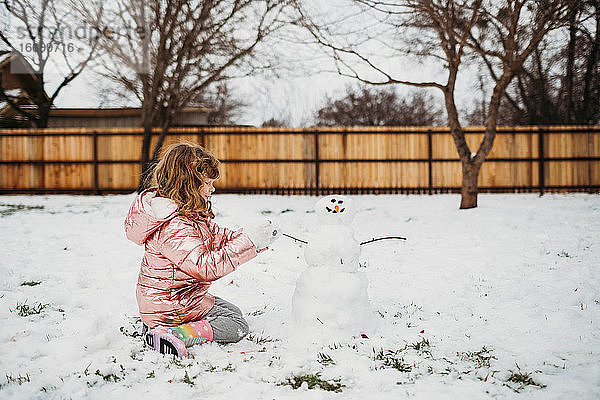 Blondes  gelocktes Mädchen sitzt draußen im Schnee und baut einen Schneemann