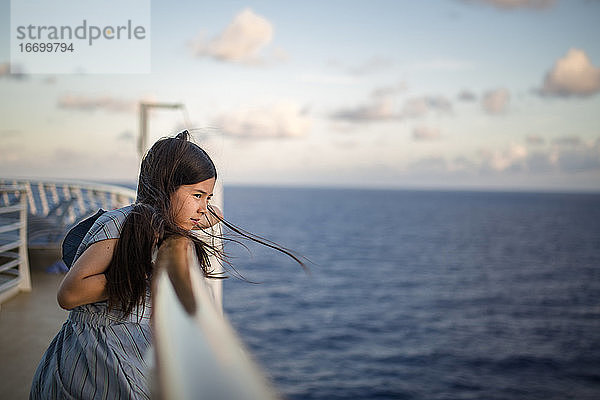 Ein junges Mädchen lehnt bei Sonnenuntergang an der Reling eines Schiffes und blickt auf das Meer