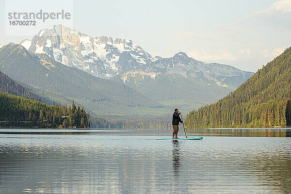 Seitenansicht eines Mannes auf einem Paddelbrett auf dem Duffey Lake vor dem Hintergrund eines spektakulären Bergrückens am Morgen in British Columbia