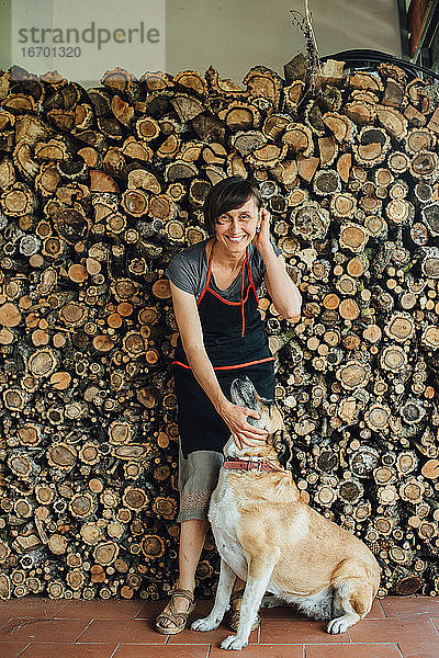 Lächelnde Frau mittleren Alters in Schürze mit Hund an einem Stapel gesägter Baumstämme