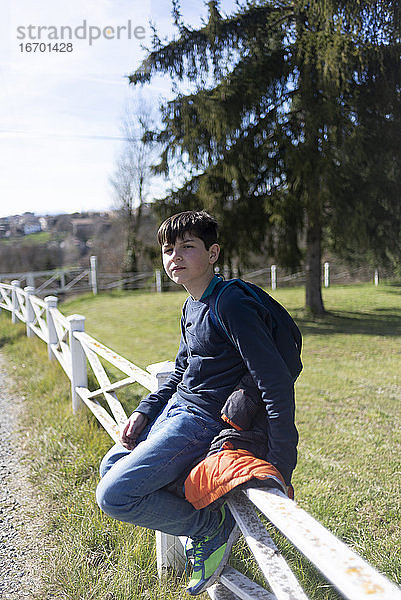 Junger Teenager  männlich  sitzt auf dem Lande am Zaun und schaut in die Kamera