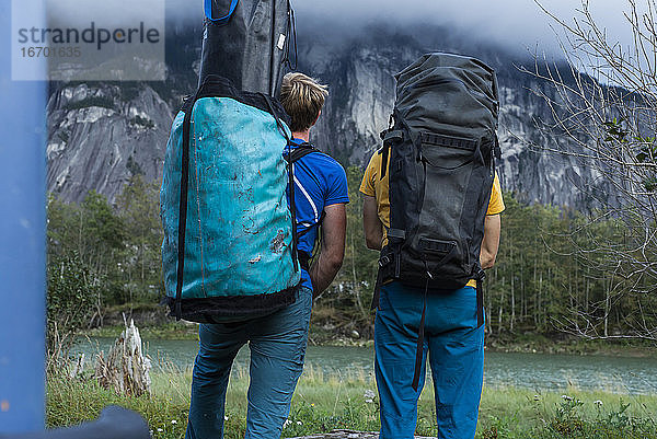 Zwei Menschen mit großem Rucksack stehen vor einem Berg und einem Fluss