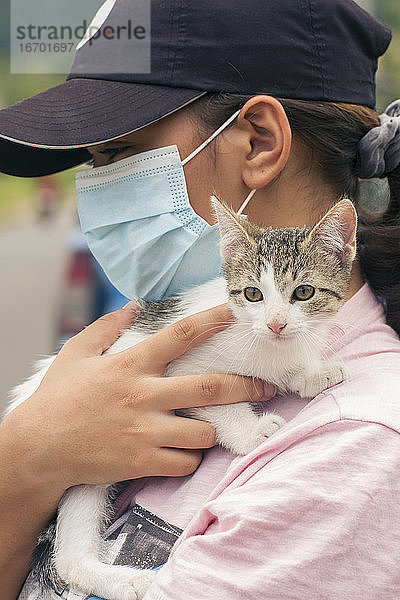 unkenntliche Frau mit Gesichtsmaske und schwarzer Mütze  die eine junge weiß-graue Katze auf den Schultern trägt  in einer verschwommenen natürlichen Umgebung