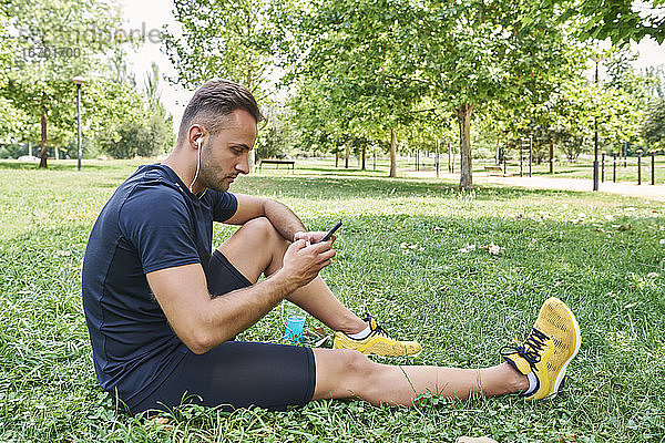 Ein Mann mit seinem Smartphone  der nach dem Training auf der Wiese eines Parks sitzt.